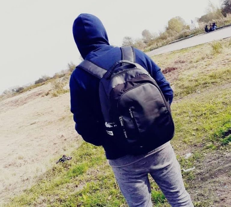 A man wearing jeans, a dark hooded jacket and backpack faces away from the camera towards a small lake and grass